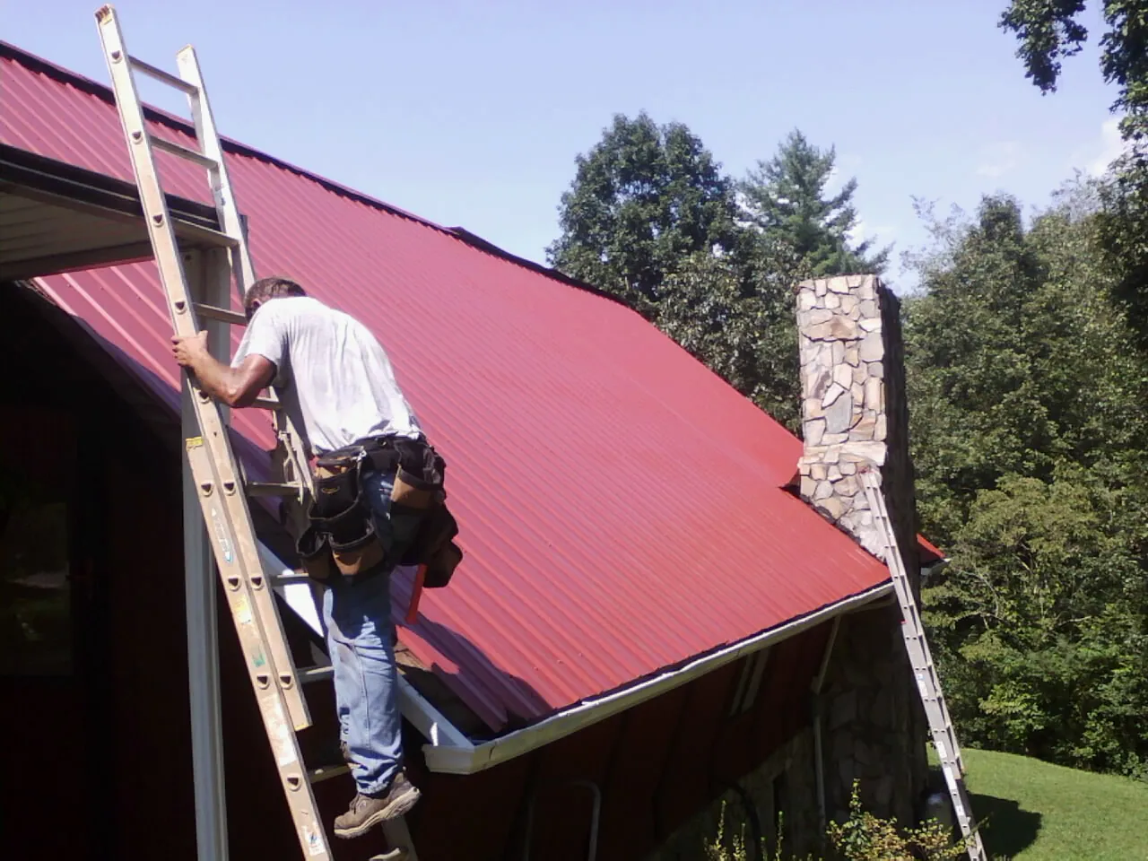 Roofer on a ladder beside a red metal roof and stone chimney — Soffit & Fascia Repair by Roofing Brothers in East Carbon